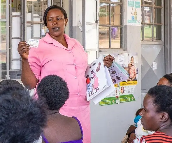A nurse in pink uniform stands in front of a row of windows, giving a family planning talk to women as they wait to immunize their children.