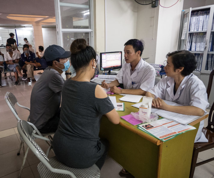 Two health workers sit at a desk strewn with papers. On the other side, two patients, their backs to the camera. Hai Duong Provincial HIV/AIDS Center, Vietnam.