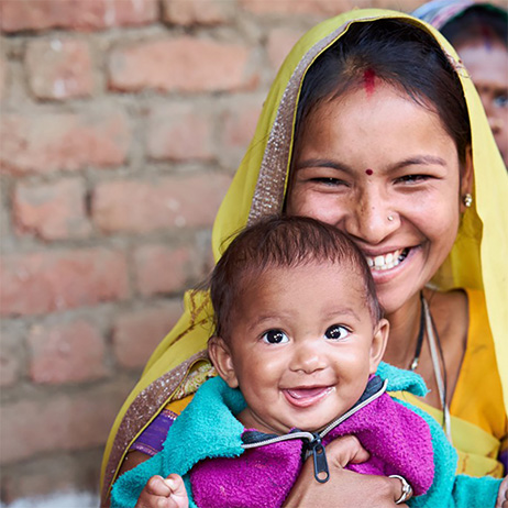 A smiling mother holds her infant who is also smiling and looking at the camera.