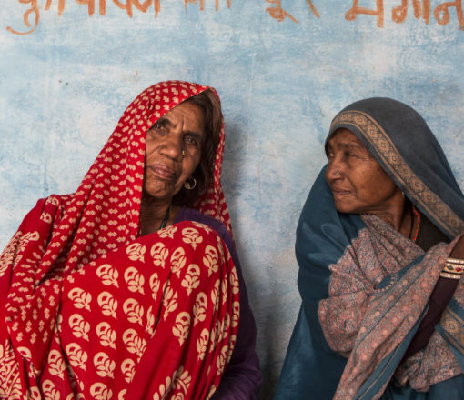 Two women sit against a blue wall in a village in Madhya Pradesh, India. Photo credit: Sujata Khanna Photography