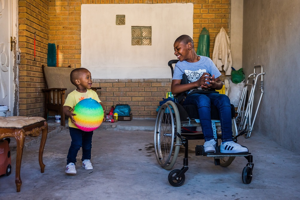 A small child holds a multi-colored ball while standing to another child on a wheelchair.